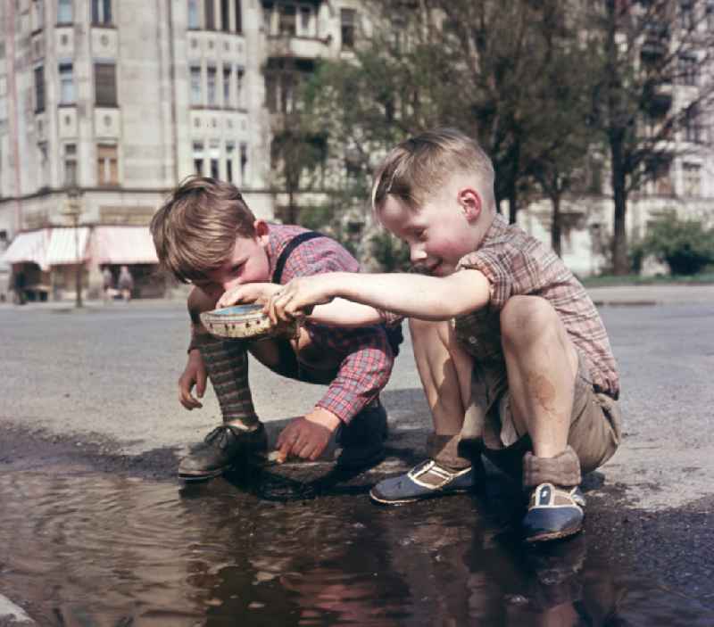 Two boys play with a toy ship at a puddle on a street in East Berlin in the territory of the former GDR, German Democratic Republic