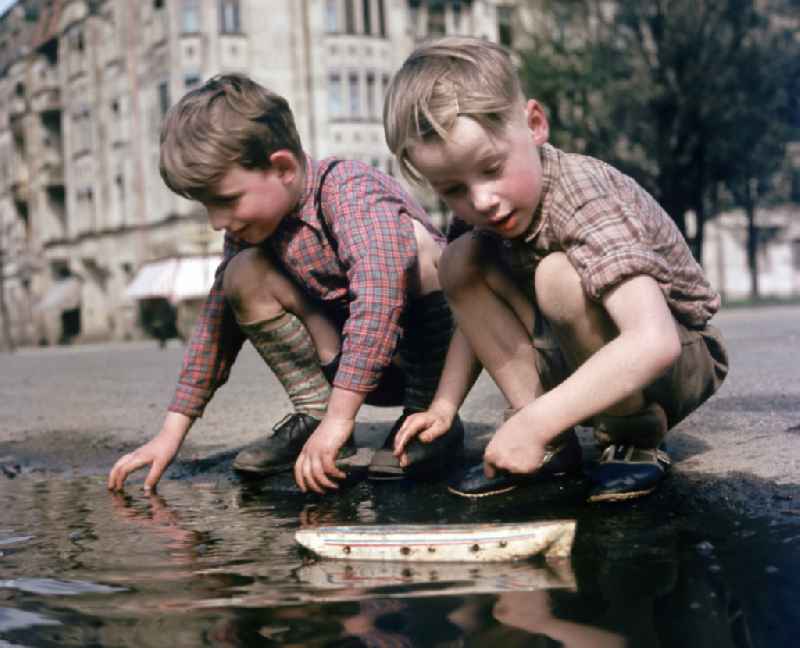 Two boys play with a toy ship at a puddle on a street in East Berlin in the territory of the former GDR, German Democratic Republic
