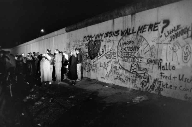 People stand together at the wall on the West Berlin side. The wall is covered in graffiti, including the words 'mom-why is this wall here?'