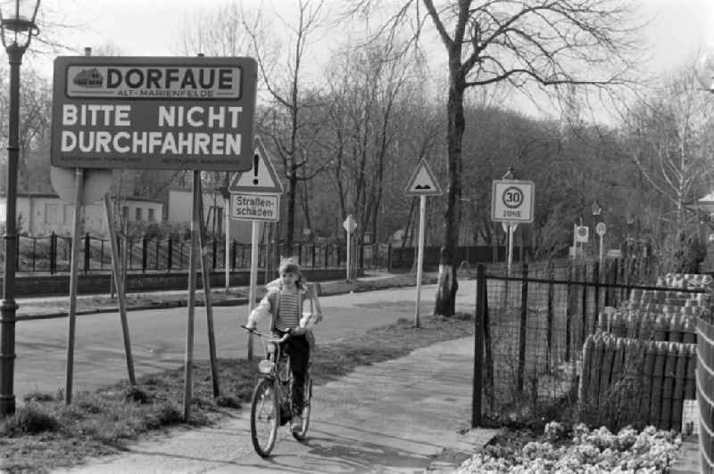 Dorfaue Buckower Chaussee in Alt-Marienfelde in Berlin. A girl rides her bike on the sidewalk from school to home, past many traffic signs and a sign saying 'Dorfaue - Please do not drive through'