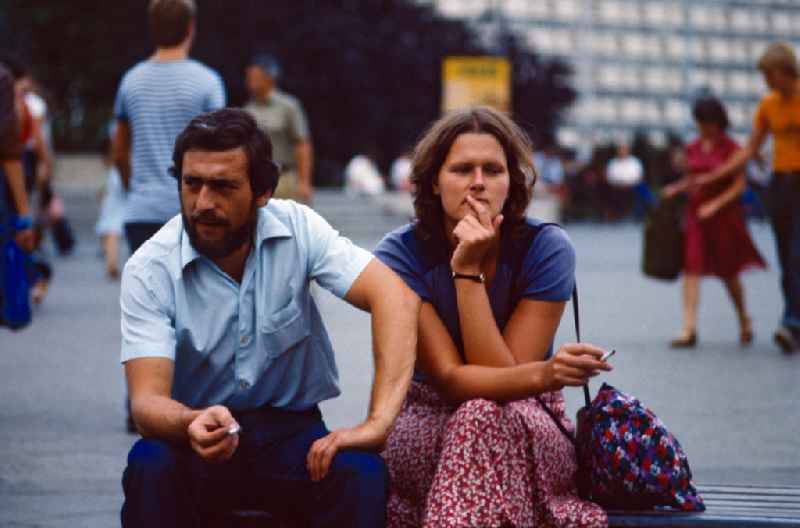 Woman and man sitting smoking on a street bench at Alexanderplatz in East Berlin in the territory of the former GDR, German Democratic Republic
