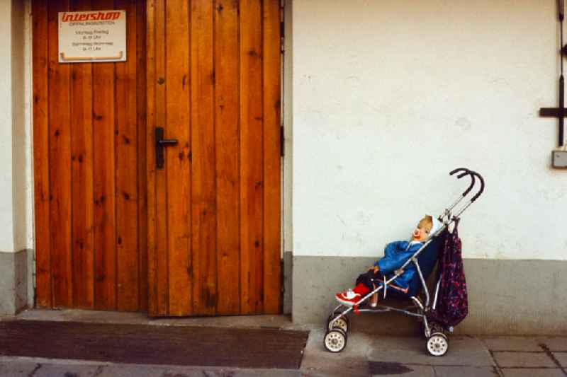 Child sits in a buggy in front of an Intershop entrance in East Berlin in the territory of the former GDR, German Democratic Republic