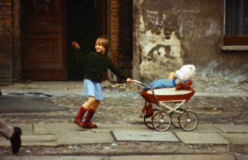 Girl pushes her teddy bear in a doll's pram on a sidewalk in East Berlin on the territory of the former GDR, German Democratic Republic