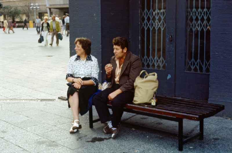 A woman and a man sit smoking on a street bench in East Berlin in the territory of the former GDR, German Democratic Republic