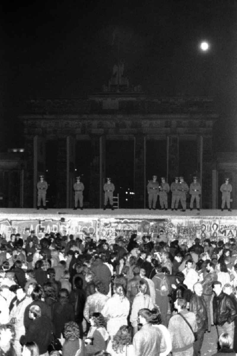 Nachtaufnahme: Blick von West-Berlin auf das Brandenburger Tor Richtung Ost-Berlin. Polizisten / Polizeibeamte / Grenzsoldaten / Soldaten stehen auf Mauer / Panzerwall / Panzermauer. Menschen / Schaulustige stehen davor. Graffiti / Schmierereien auf Mauer.