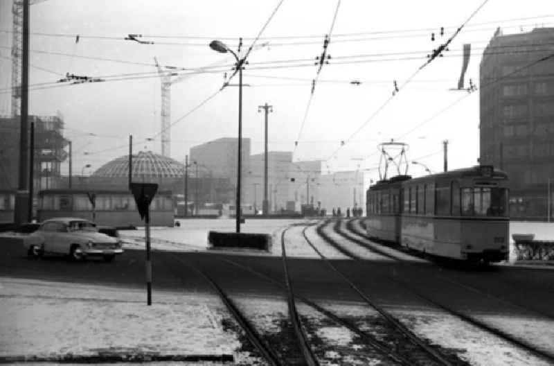 Alexanderplatz verschneit, mit Baustelle Congresshalle und Haus des Lehrers im Hintergrund. Auto steht auf Straße und läßt Straßenbahn vorbeifahren. Schienen auf Straße.