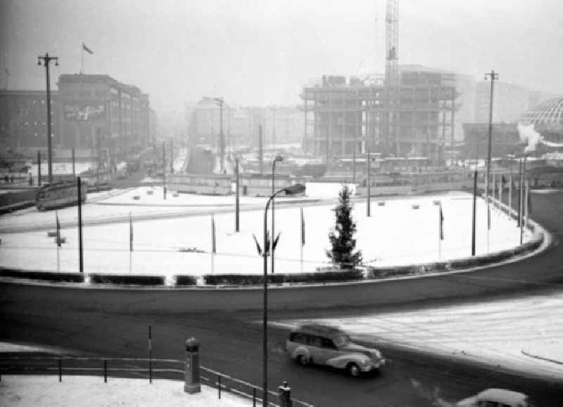 Verschneiter Kreisverkehr auf dem Alexanderplatz. Auto fährt auf Straße aus Kreisverkehr / biegt ab. Baustelle Haus des Lehrers mit Kran im Hintergrund.