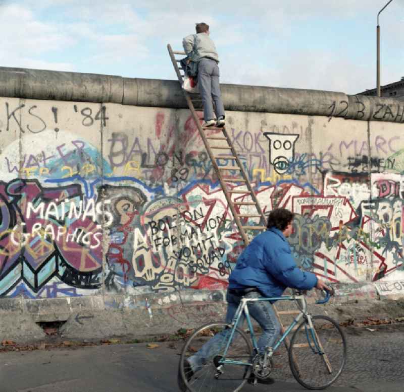 Neugieriger Mann klettert mit Hilfe einer Leiter an der Berliner Mauer hoch. Die Berliner Mauer war während der Teilung Deutschlands ein hermetisch abriegelndes Grenzbefestigungssystem der Deutschen Demokratischen Republik.