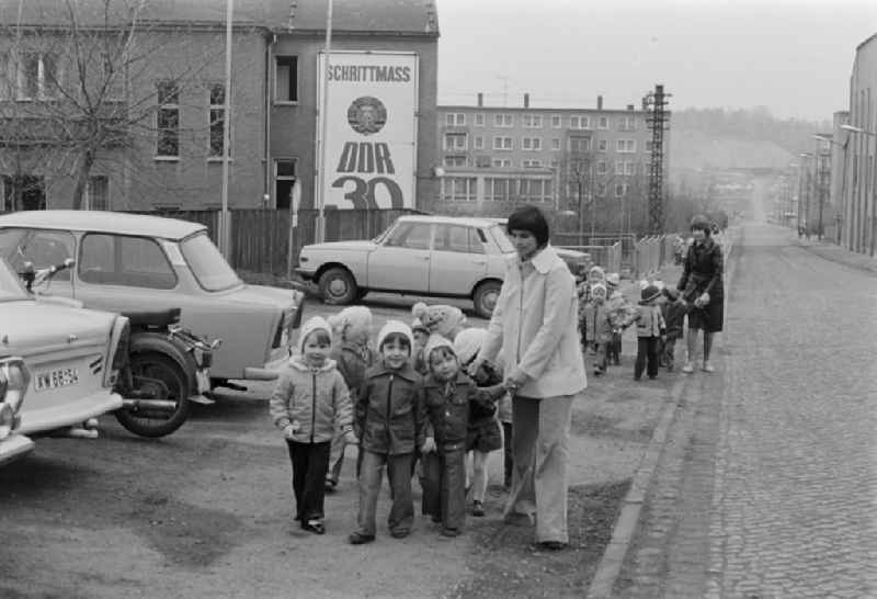 Educators look after small children in groups on the sidewalk of a street in front of the background of a propaganda poster 'Schrittmass DDR 3