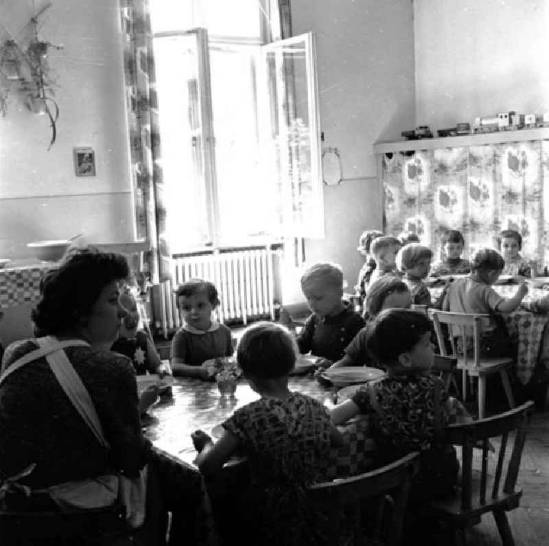 Kinder sitzen zusammen am Tisch beim Mittagessen im Kindergarten, Erzieherin sitzt daneben.