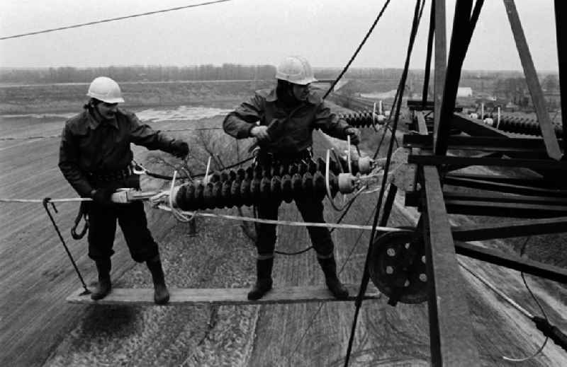 Arbeiter bei der Montage einer Hochspannungsleitung bei Brieselang in luftiger Höhe in Aktion. Die DDR-Energieversorung basiert zu einem großen Teil auf der Verstromung von Braunkohle aus der Lausitz und Sachsen, was unter anderem lange Leitungswege nötig macht.