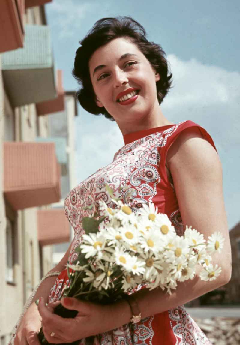 A young woman poses with flowers in front of a newly built block of flats in Coswig, Saxony in the territory of the former GDR, German Democratic Republic