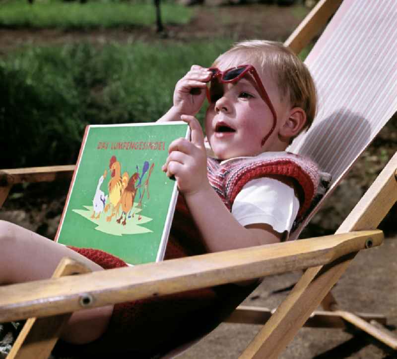 A boy sits on a deck chair with a book and sunglasses in Coswig, Saxony in the territory of the former GDR, German Democratic Republic