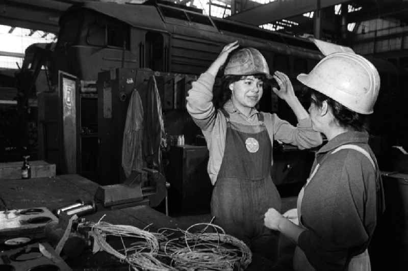 Zwei junge Damen im RAW (Reichsbahnausbesserungswerk) Cottbus stehen zusammen und setzen zum Arbeitsschutz eine Helm auf. Im Rahmen des polytechnischen Unterricht nahmen die Schüler aktiv an der DDR-Produktion teil.