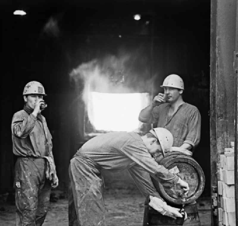 Arbeiter vor einem Ofen im Eisenhüttenkombinat Ost (EKO) zapfen aus einem Fass Getränke.