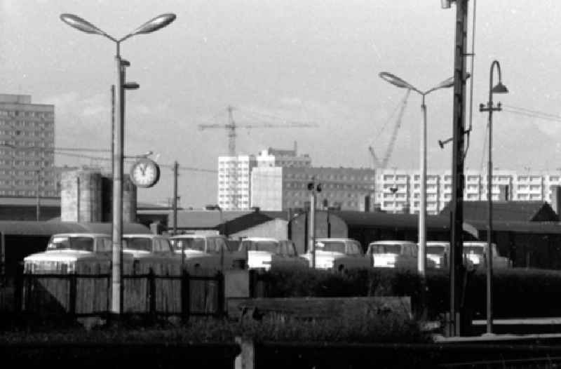 Blick über Autos der Marke Moskwitsch 412 auf auf Schienenwaggon und Güterbahnhof auf Plattenbauten mit Kränen. Bahnhofsuhr steht auf kurz nach elf, daneben Laterne / Straßenlampe.