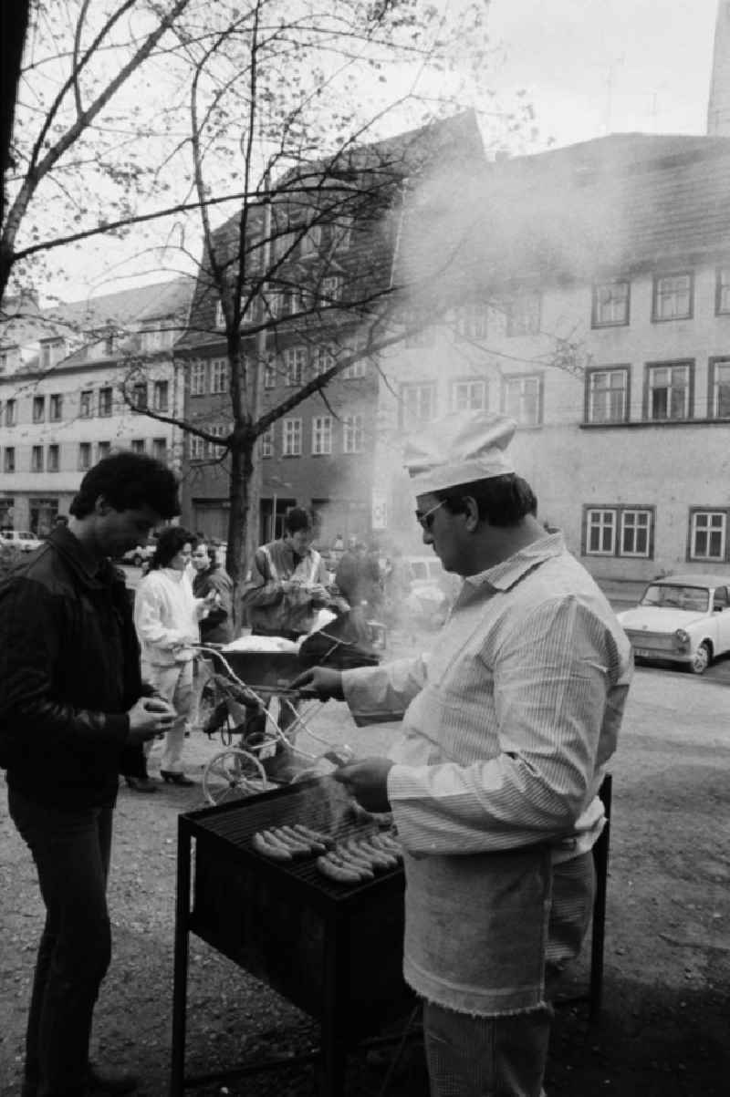 Unweit der berühmten Krämerbrücke in Erfurt werden Rostbratwürste verkauft. Koch mit Kochmütze und Kochkleidung steht vor einem Grill mit Bratwürsten darauf. Im Hintergrund parkt ein Auto vom Typ Trabant 601. Die sogenannten Roster sind eine Spezialität aus Thüringen, die erstmals 14