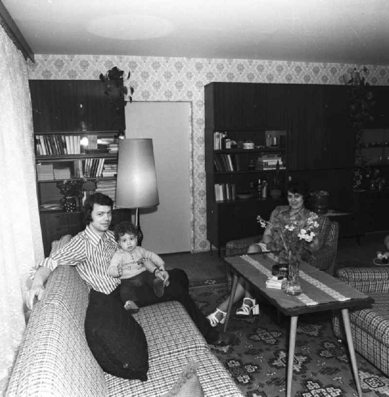 Wohnzimmer in der Neubauwohnung einer jungen Familie im Stil der siebziger Jahre. Im Hintergrund Schrankwand Typ Hellerau. Paar mit Kind auf dem Sofa / der Couch.
