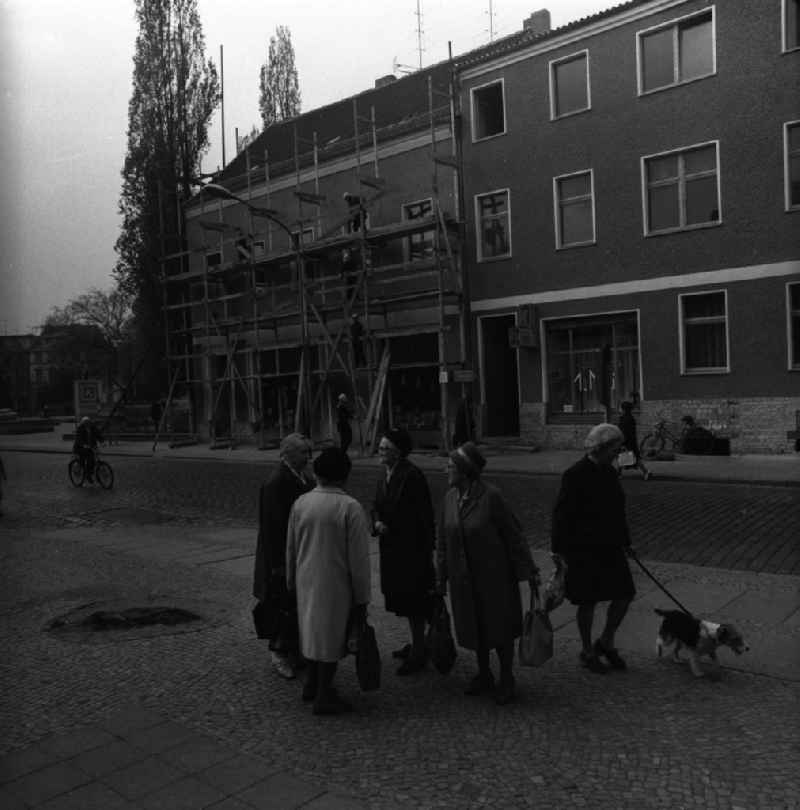 Gruppe älterer Damen führt eine Unterhaltung auf dem Bürgersteig. Passanten nähern sich der Gruppe, gehen an ihr vorbei und überqueren die Straße. Im Hintergrund bauen Arbeiter ein Gerüst ab.