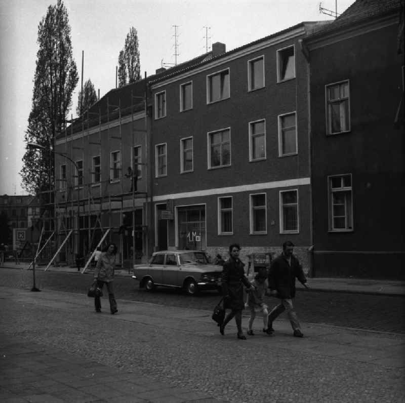 Alltagsszene / Stadtansicht. Auf dem Bürgersteig eine Familie und eine Fußgängerin, im Hintergrund demontieren Bauarbeiter ein Baugerüst. Auf der Straße ein russischer PKW Typ Moskwitsch 412.