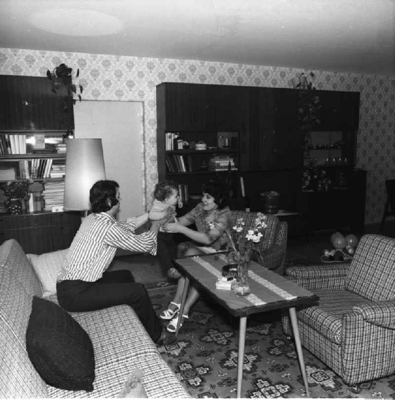 In der Wohnung einer jungen Familie / im Wohnzimmer. Die Wohnung ist im Stil der siebziger Jahre eingerichtet. Im Hintergrund Schrankwand Typ Hellerau. Das Paar sitzt mit seinem Kind / Baby auf dem Sofa.