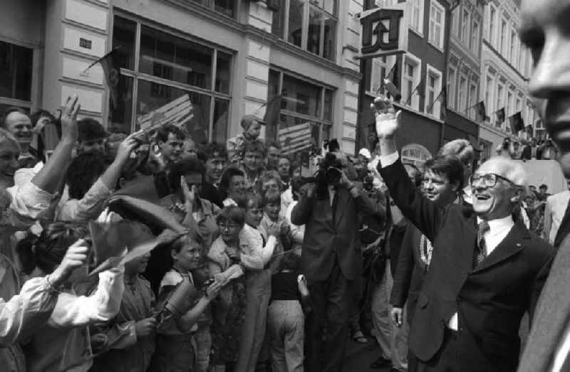 Besuch von Erich Honecker anlässlich der Domweihe. Passanten / Schaulustige auf Marktplatz, stehen hinter Absperrung und geben Applaus. Honecker steht davor lacht und winkt, dahinter Klaus Ewald, Oberbürgermeister Greifswald. DDR-Flaggen hängen an Gebäuden.