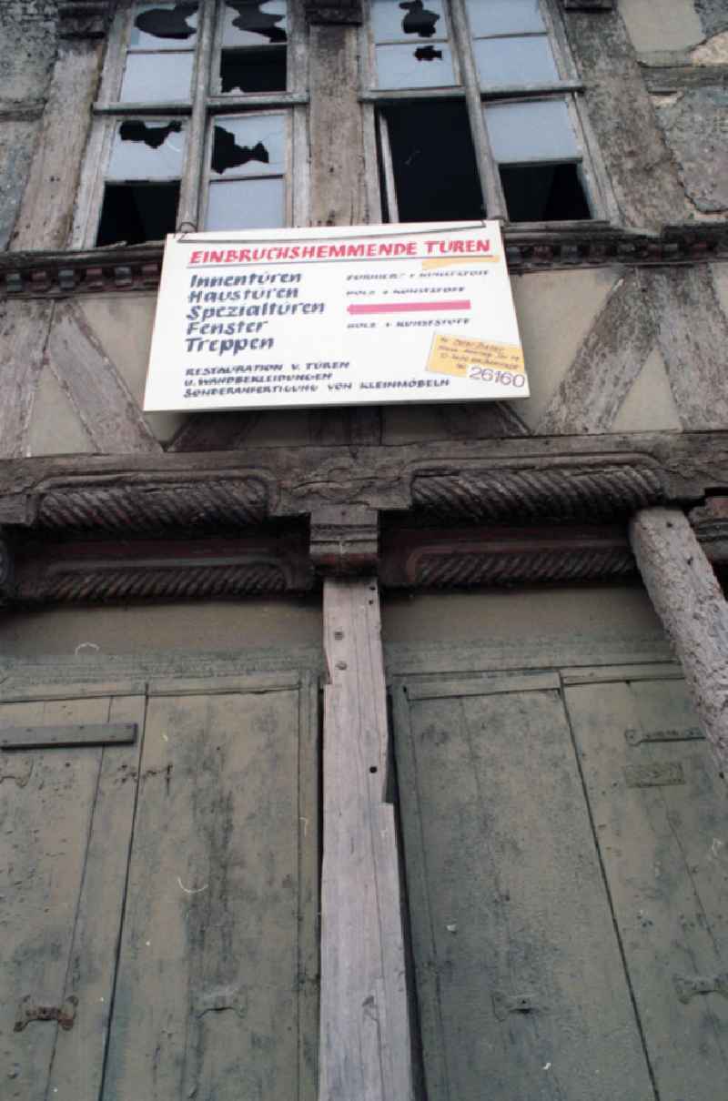 Werbeschild über einer Haustür eines stark sanierungsbedürftigen Fachwerkhaus, mit der Aufschrift 'Einbruchshemmende Türen ...', darüber Fenster mit zerbrochenen / kaputten Fensterscheiben.