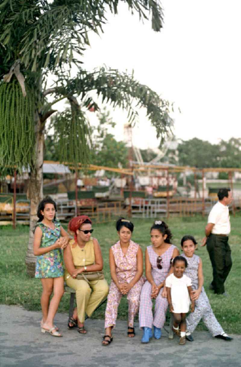 Frauen und Kinder sitzen auf einer Bank vor einem kleinen Rummel in Havanna.