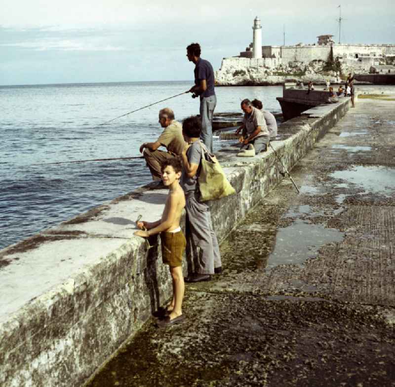 Angler sitzen auf der Mauer an der acht Kilometer langen Uferpromenade, dem sogenannten Malecón in Havanna. Im Hintergrund die historische Festung 'El Morro', eines der Wahrzeichen der kubanischen Hauptstadt.