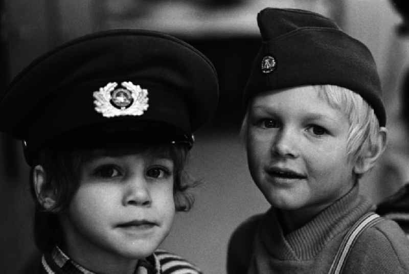 Kinder in der Kinderkombination / im Kindergarten 'Pünktchen' in Hennigsdorf tragen  Armee-Kopfbedeckungen / NVA Mützen. 98 Prozent aller Drei- bis Sechsjährigen war in der DDR ein Kindergartenplatz sicher. Die Öffnungszeiten wurden an die Arbeitszeiten der werktätigen Mütter angepasst, damit Frauen gleichberechtigter Teil der Gesellschaft sein konnten. In den Plänen war auch vorgesehen, dass die Kinder 'freundschaftliche Gefühle zu den Soldaten der NVA herausbilden'.