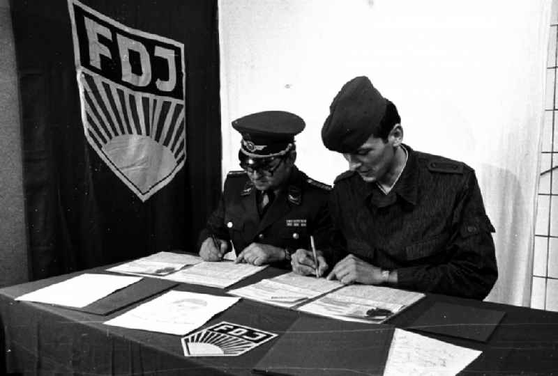 Soldier and ensign in the uniform of the LSK/LV Air Force of the FTB-9 Aviation Technical Battalion of the NVA in front of an FDJ flag at a table in Karlshagen, Mecklenburg-Western Pomerania in the area of the former GDR, German Democratic Republic