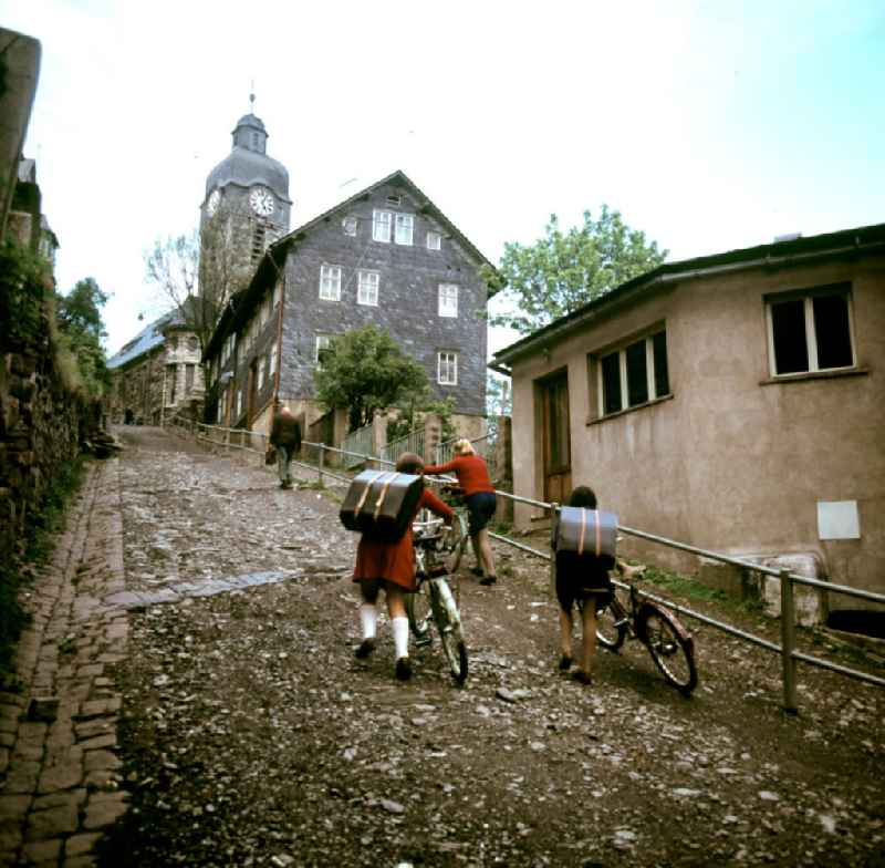 Zwei Mädchen mit Akkordeon schieben ihre Fahrräder den Berg hoch in Lauscha im Thüringer Wald. Im Hintergrund die Lauschaer Kirche. Die Fassaden und Dächer der Häuser des Ortes sind mit Schiefer bedeckt. Der Thüringer Wald mit seinen Wander- und Erholungsmöglichkeiten war ein beliebtes Urlaubsziel in der DDR.