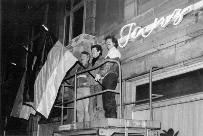 Demonstranten stehen zusammen auf einen Gerüst vor Flaggen der BRD. Dahinter ein Leuchtreklame mit der Aufschrift 'Tanz'. Der Wunsch nach einem wiedervereinigten Deutschland bestimmte die Leipziger Montagsdemonstration. Die politische Eigenständigkeit der DDR wurde nur noch auf wenigen Transparenten gefordert.