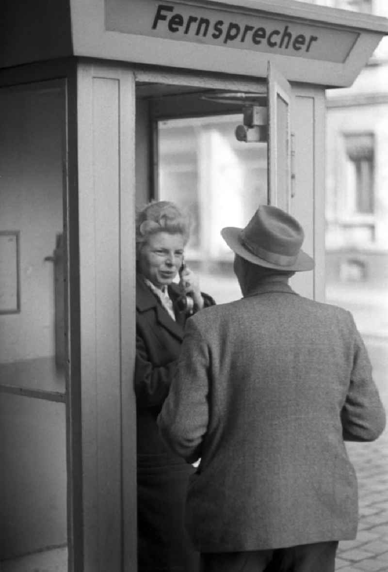 Eine Frau telefoniert in einem Telefonhäuschen in Leipzig mit einem öffentlichen Fernsprecher, ihr Mann hört an der geöffneten Tür mit zu.