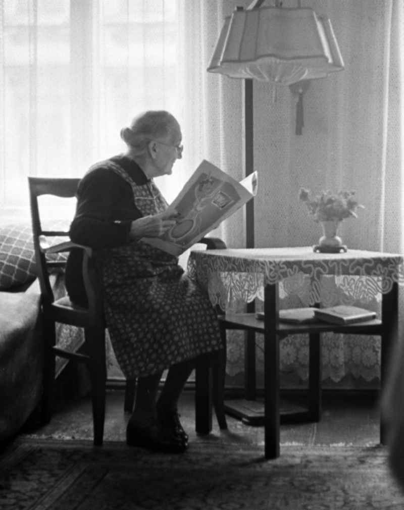 Eine alte Frau sitzt in ihrem Zimmer mit typischer Möblierung und liest interessiert die Zeitung.