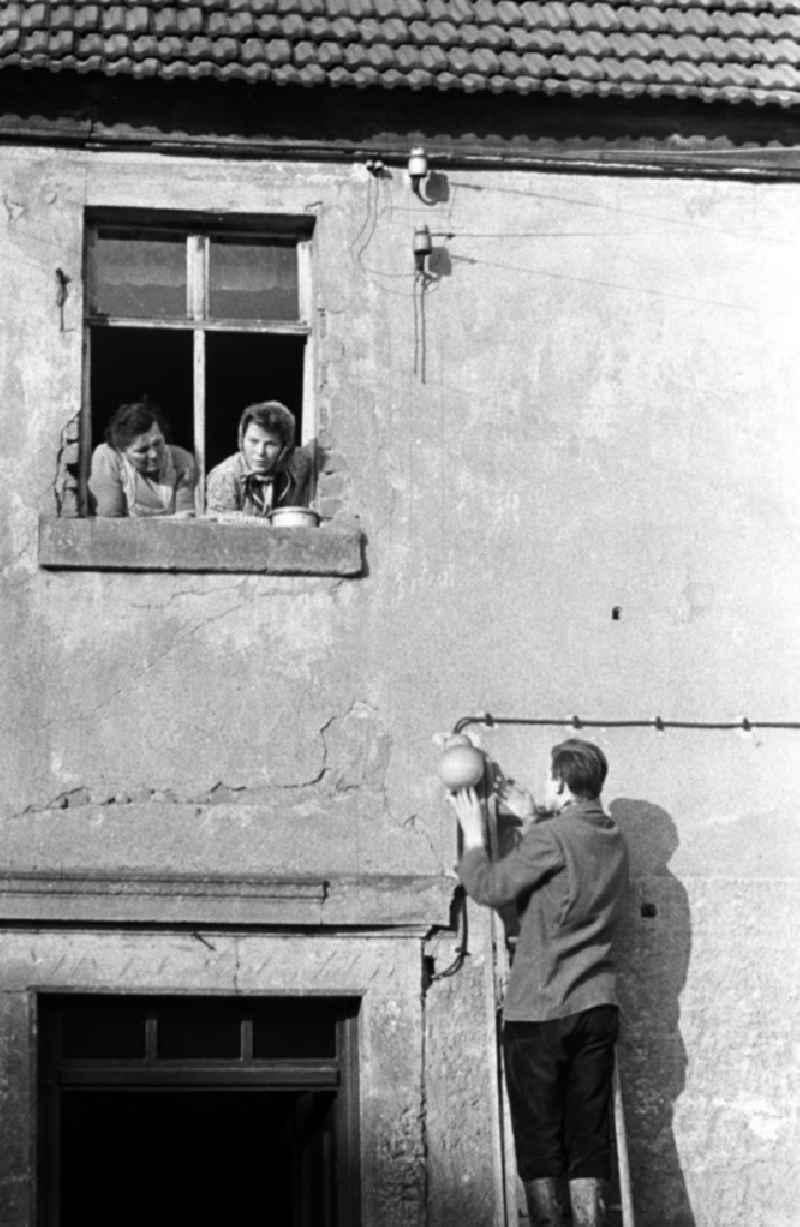 Ein Mann steht an einer Hauswand auf einer Leiter und repariert eine Lampe, zwei Frauen schauen ihm aus dem Fenster heraus dabei zu.
