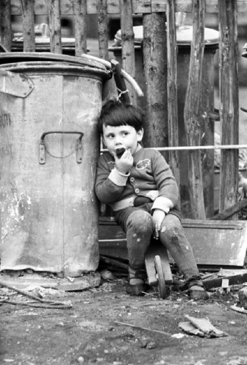 Ein Kind sitzt auf seinem Holzroller in einer dreckigen Ecke neben einer alten Mülltonne. Den Jungen scheint der Müll nicht zu stören - genüßlich beißt er in ein Stück Kuchen.