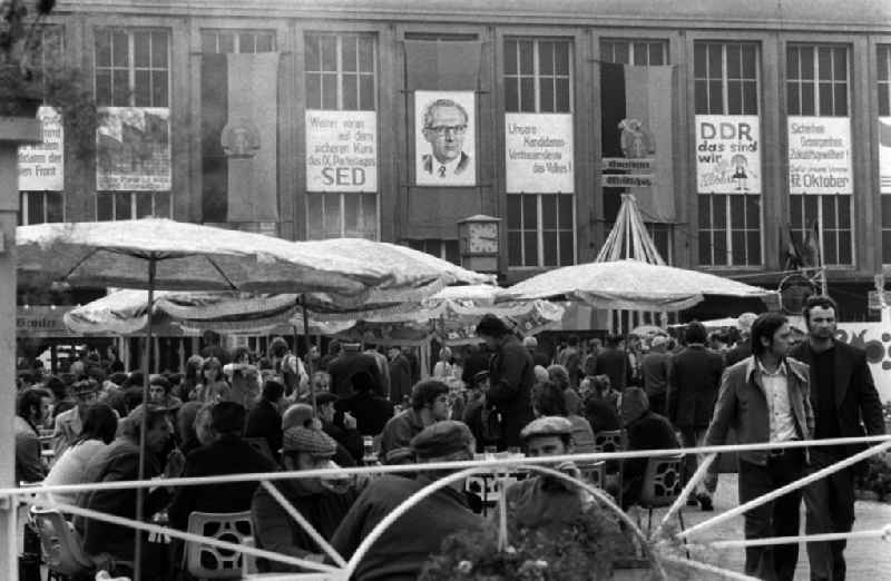 Blick auf Passanten sitzen an Tischen unter Sonnenschirmen beim Essen und Trinken auf den gut besuchten Konsum Markt in Merseburg. Im Hintergrund sind Transparente mit politischen Parolen sowie ein Porträt von DDR Staatschef und Parteichef Erich Honecker zu sehen.