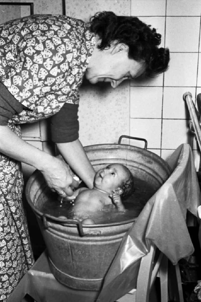 A woman in smock apron bathes a baby in a zinc tub in Merseburg in the federal state Saxony-Anhalt in the area of the former GDR, German democratic republic