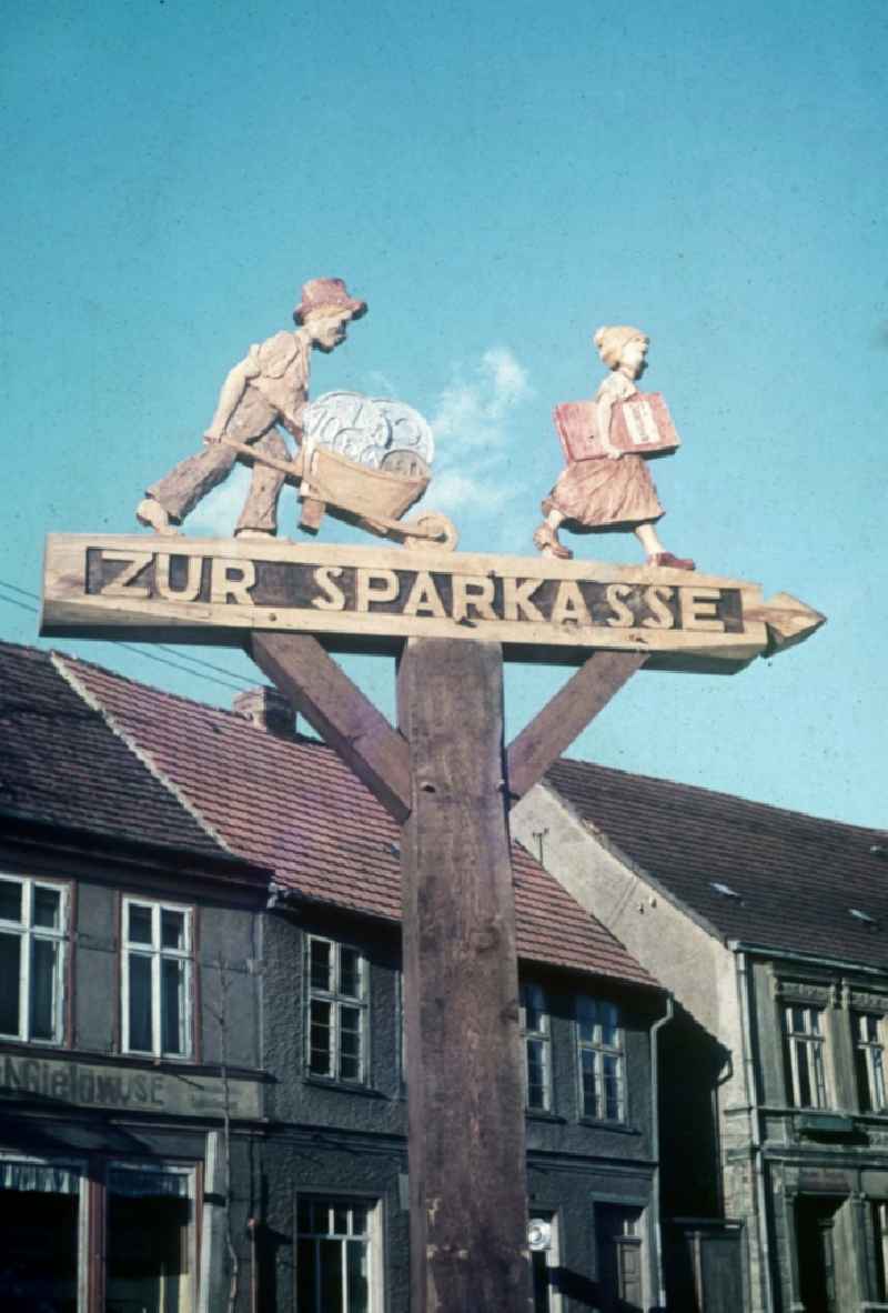 A carved signpost with figures of wood him the way to the savings bank points in Neustrelitz in the federal state Mecklenburg-West Pomerania in the area of the former GDR, German democratic republic