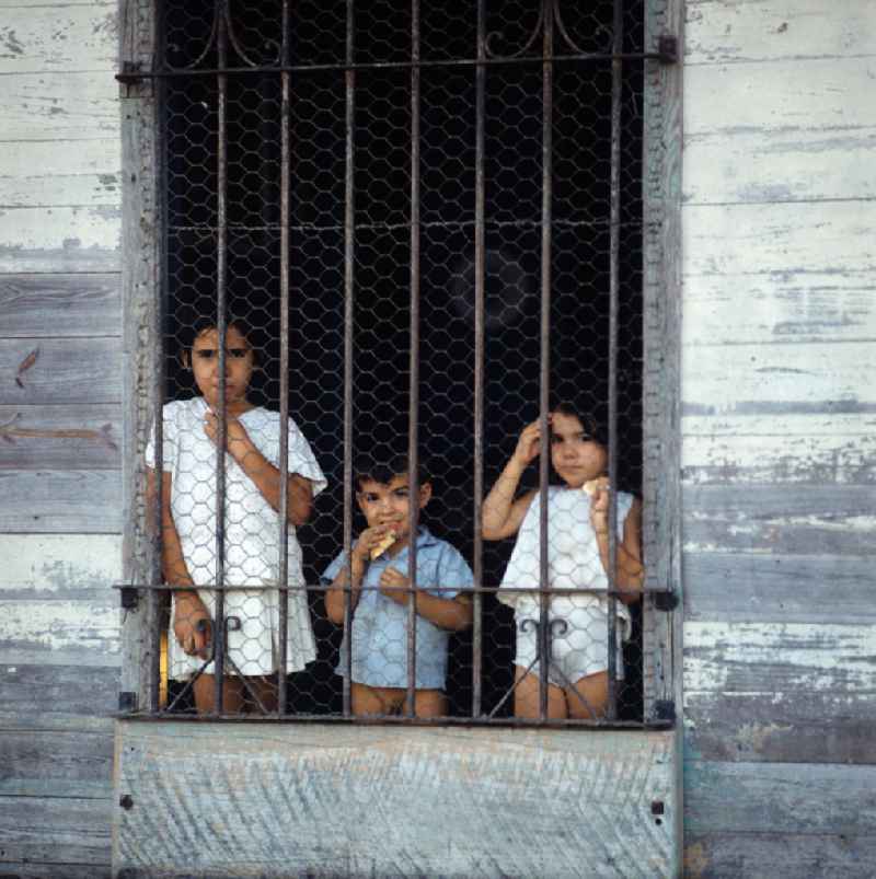 Drei Kinder blicken neugierig durch ein vergittertes Fenster in einem Haus in der kubanischen Hafenstadt Nuevitas. Children at the window in the port city Nuevitas.