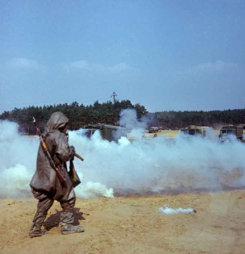 Manöverübung der NVA-Truppen der chemischen Abwehr (ABC-Abwehr) in Peenemünde. Soldat mit Schutzanzug (ABC-Anzug, Gummianzug), Gasmaske und Waffen kurz vor dem Durchqueren einer Qualmwand. Im Hintergrund Armee-LKWs.