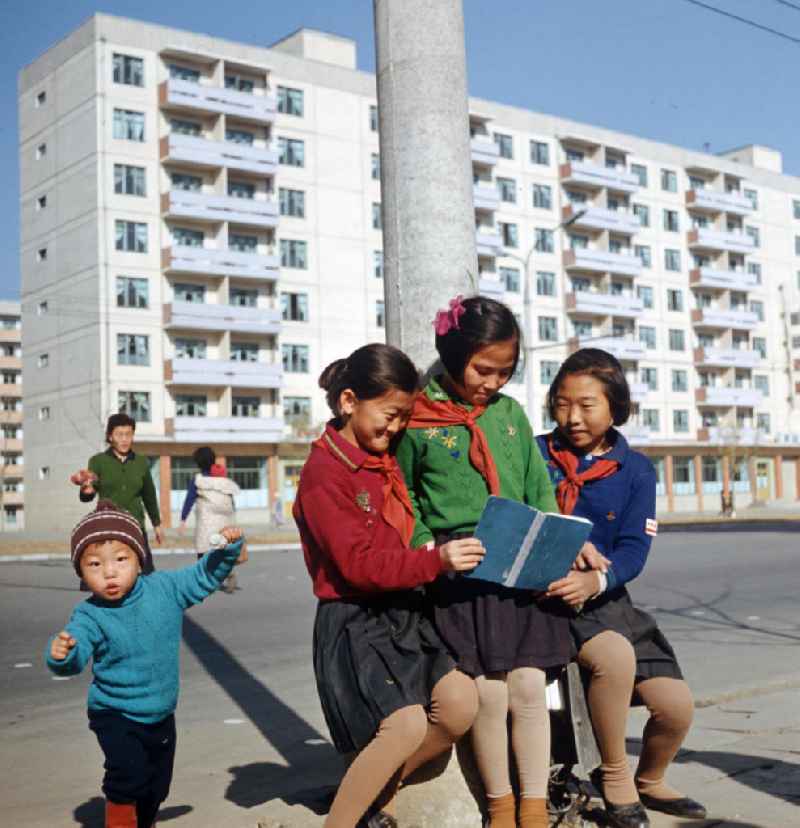 Mädchen in Schuluniform mit rotem Halstuch schauen gemeinsam ein Buch an auf einer Straße in Pjöngjang, der Hauptstadt der Koreanischen Demokratischen Volksrepublik (KDVR). Im Hintergrund Plattenbauten im sozialistischen Stil. Die nordkoreanische Hauptstadt war nach der Zerstörung im Koreakrieg vor allem mit Unterstützung der Sowjetunion in den 50er und 6