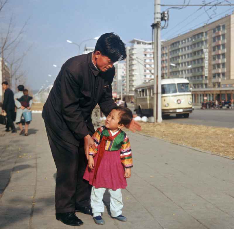 Mann mit Kind auf einer Straße in Pjöngjang, der Hauptstadt der Koreanischen Demokratischen Volksrepublik (KDVR). Im Hintergrund Plattenbauten im sozialistischen Stil. Die nordkoreanische Hauptstadt war nach der Zerstörung im Koreakrieg vor allem mit Unterstützung der Sowjetunion in den 50er und 6
