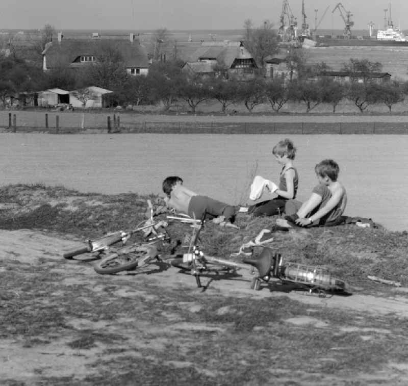 Children who have taken off their bicycles are sitting at the edge of the field in Rostock in the federal state Mecklenburg-Western Pomerania on the territory of the former GDR, German Democratic Republic. In the background the overseas port of Rostock