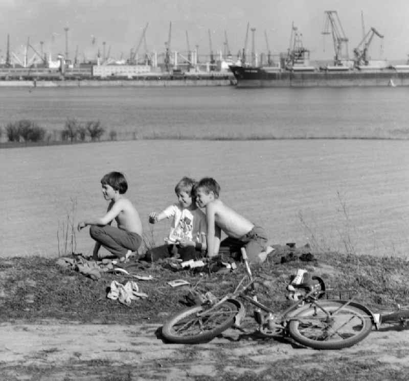 Children who have taken off their bicycles are sitting at the edge of the field in Rostock in the federal state Mecklenburg-Western Pomerania on the territory of the former GDR, German Democratic Republic. In the background the overseas port of Rostock