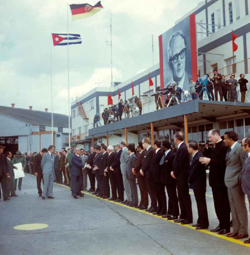 Der Staats- und Parteivorsitzende der DDR, Erich Honecker, wird auf dem Flughafen Santiago de Cuba von dem kubanischen Regierungschef Fidel Castro und weiteren Regierungsvertretern in Empfang genommen. Im Hintergrund Vertreter der Presse und ein großes Abbild des Staatsgastes. Honecker stattete vom 2
