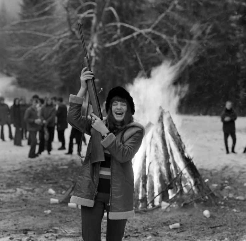 A woman holds up an air rifle while a campfire burns in the background. She is a member of the Polish Landjugend, which is on a trip to Schmiedefeld am Rennsteig on the territory of the former GDR, German Democratic Republic