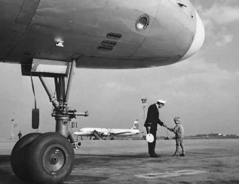 A marshaller the INTERFLUG talking to a boy on the tarmac at the airport at Schoenefeld in what is now the state of Brandenburg. In the foreground the suspension of IL-18. In the background is a propeller aircraft the airline INTERFLUG with gangway
