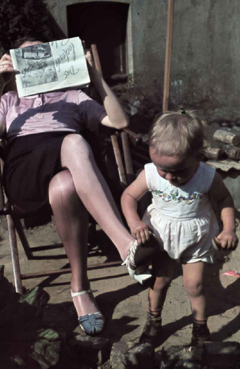 Kleinkind spielt im Garten, die Mutter sitzt im Liegestuhl und liest eine Zeitung. Plying infant in a garden. The mother is sitting in a deck chair and read a newspaper.